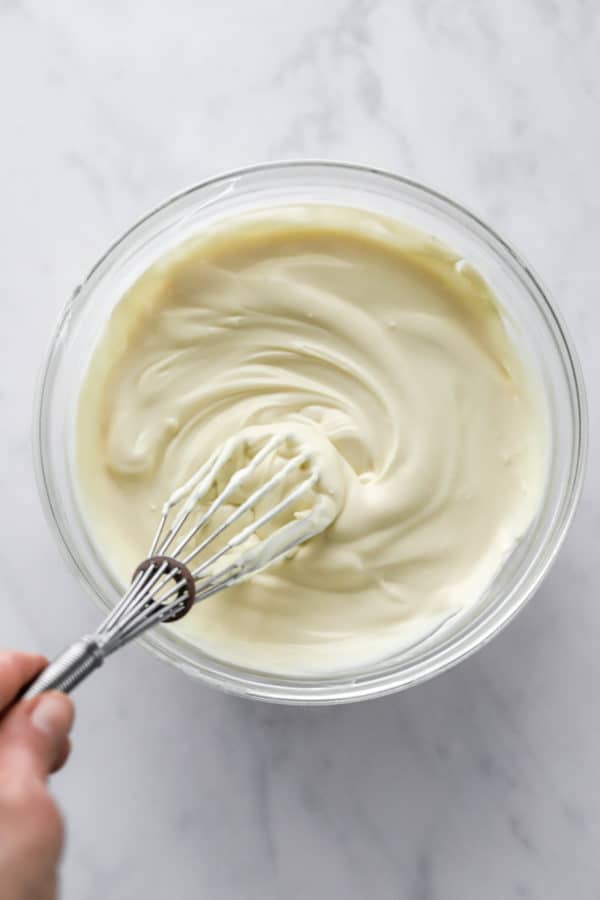 Hand using a metal whisk to mix vanilla frosting in a glass bowl.