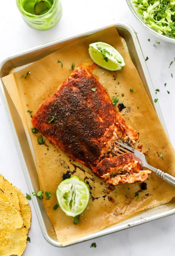 Cooked, charred salmon on a baking sheet with a fork breaking off of a piece with limes on the pan with the salmon.
