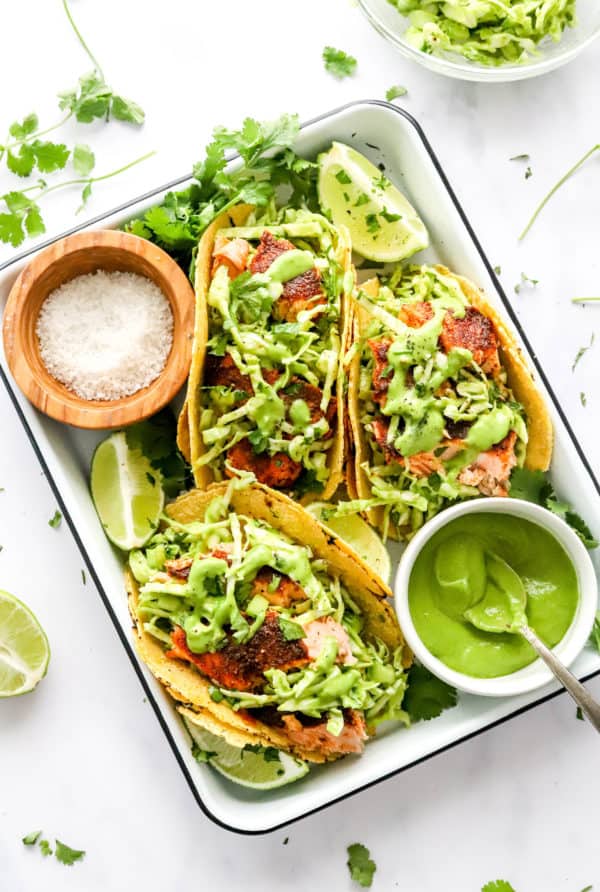 Three salmon tacos on a white pan with a bowl of salt behind them and a bowl of green dressing in front of them with green salad on top of the tacos.