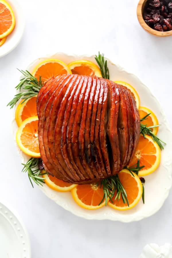 Cooked ham with glaze on it on a white platter with orange slices and rosemary around it.