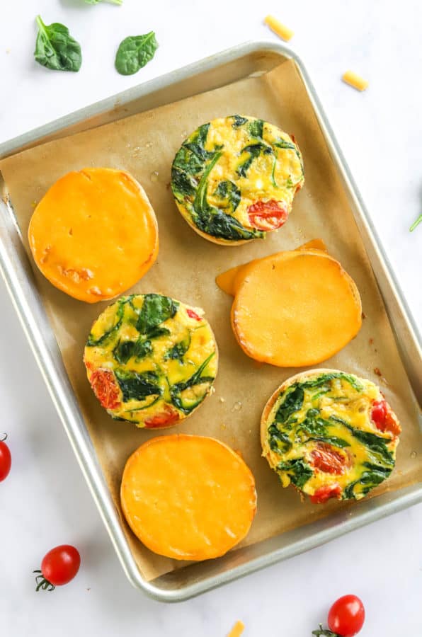 6 English muffins topped with melted cheese and egg and veggies circles on a baking sheet.