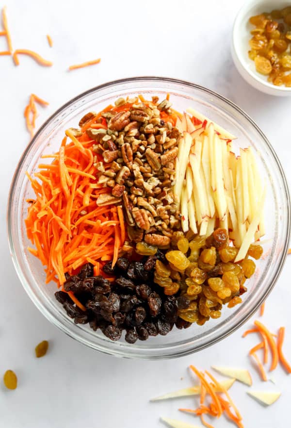Shredded apple and carrot, raisins and pecans in a round glass bowl.