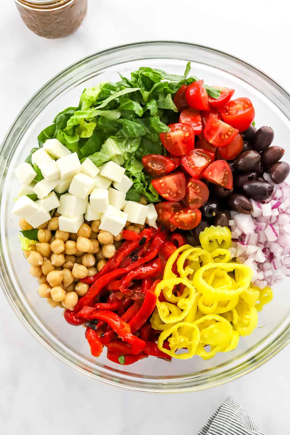 Sliced romaine lettuce, sliced tomatoes, cubed cheese, chickpeas, peppers and onions in a glass salad bowl. 
