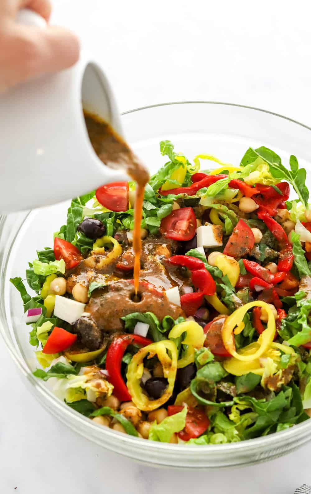 Dressing being poured onto a salad with greens, peppers and olives in it in a glass serving bowl. 