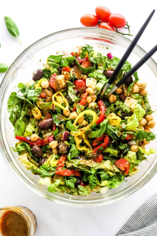 Colorful green salad in a salad bowl with Italian ingredients on it. With black serving spoons sticking out of the bowl.