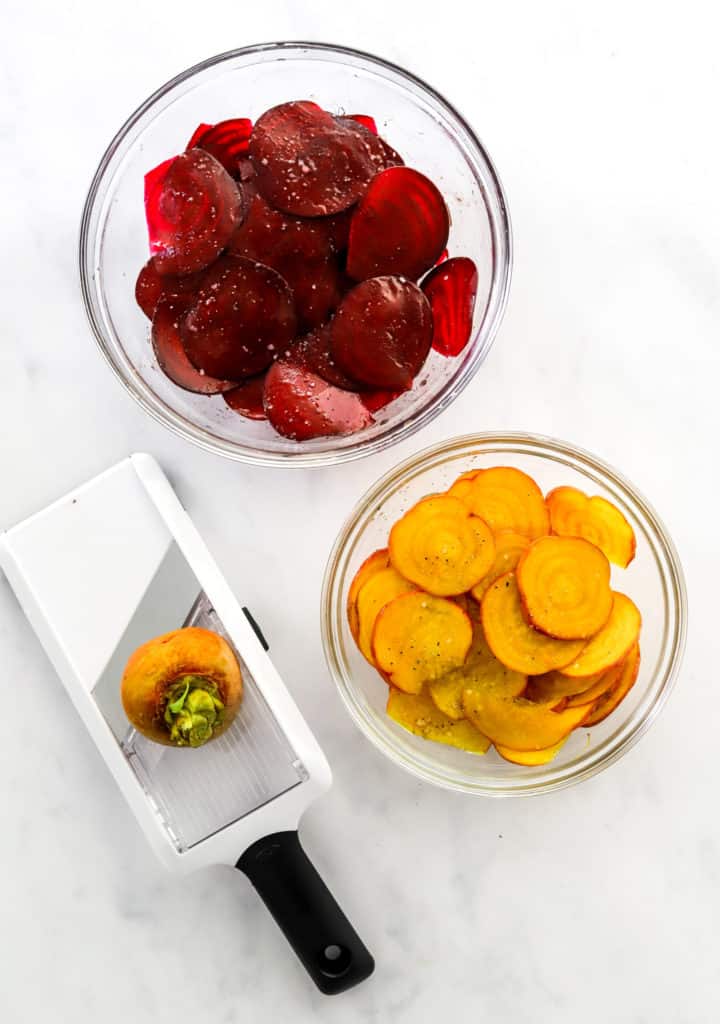 Glass bowl of sliced red beets with another glass bowl of sliced yellow beets in front of it with a mandolin with a yellow beet on it in front of it.
