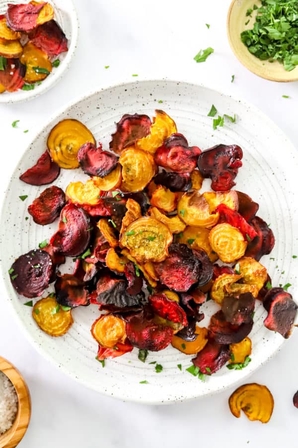 Pile of baked red and golden beet chips on a white and brown specked plate with more behind it.