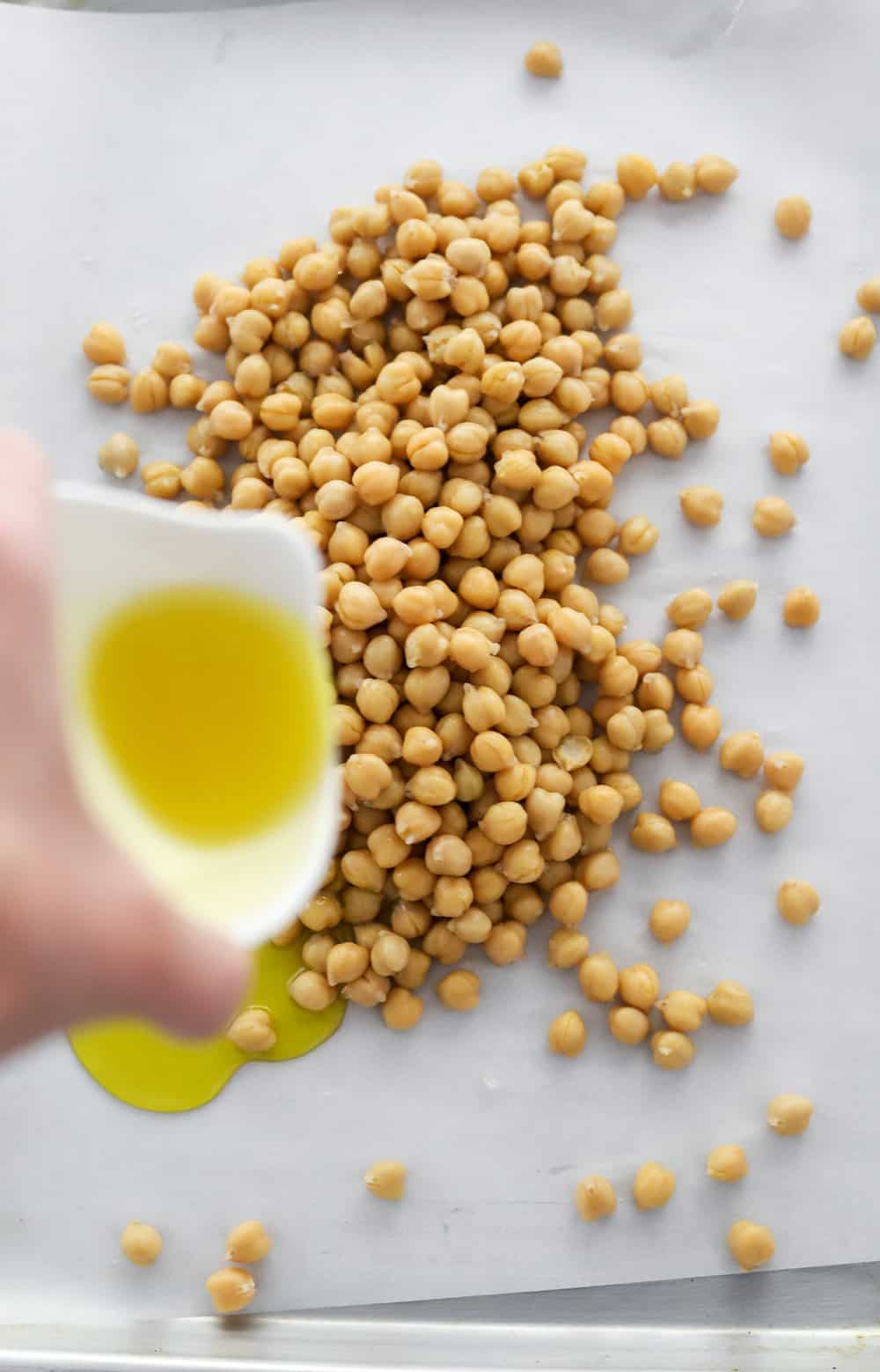 Hand pouring olive oil over chickpeas on a baking sheet