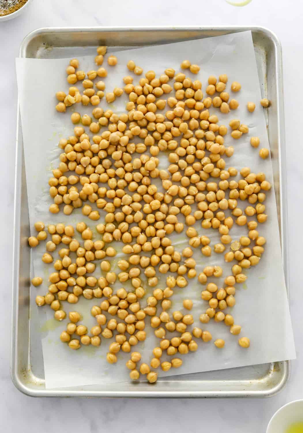 Uncooked chickpeas covered in olive oil on a baking sheet lined with white parchment paper.