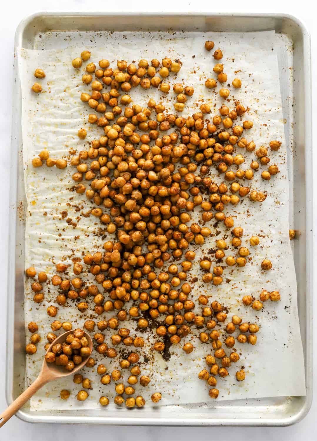 Crispy, seasoned chickpeas that are golden brown on a parchment paper lined baking sheet with a small wooden spoon on the pan. 