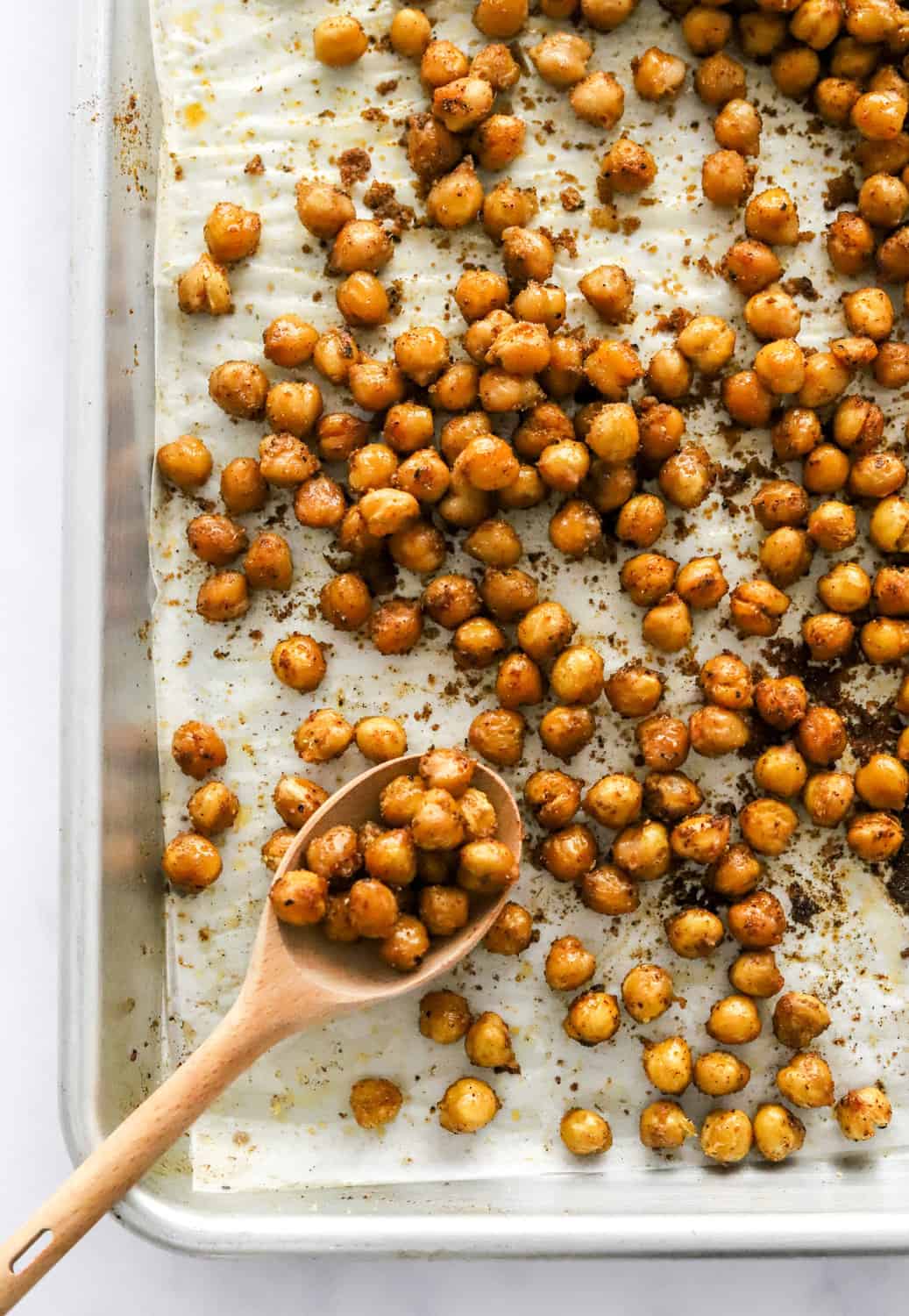 Oven roasted chickpeas on a baking sheet with a wooden spoon with some chickpeas on it on the baking sheet. 