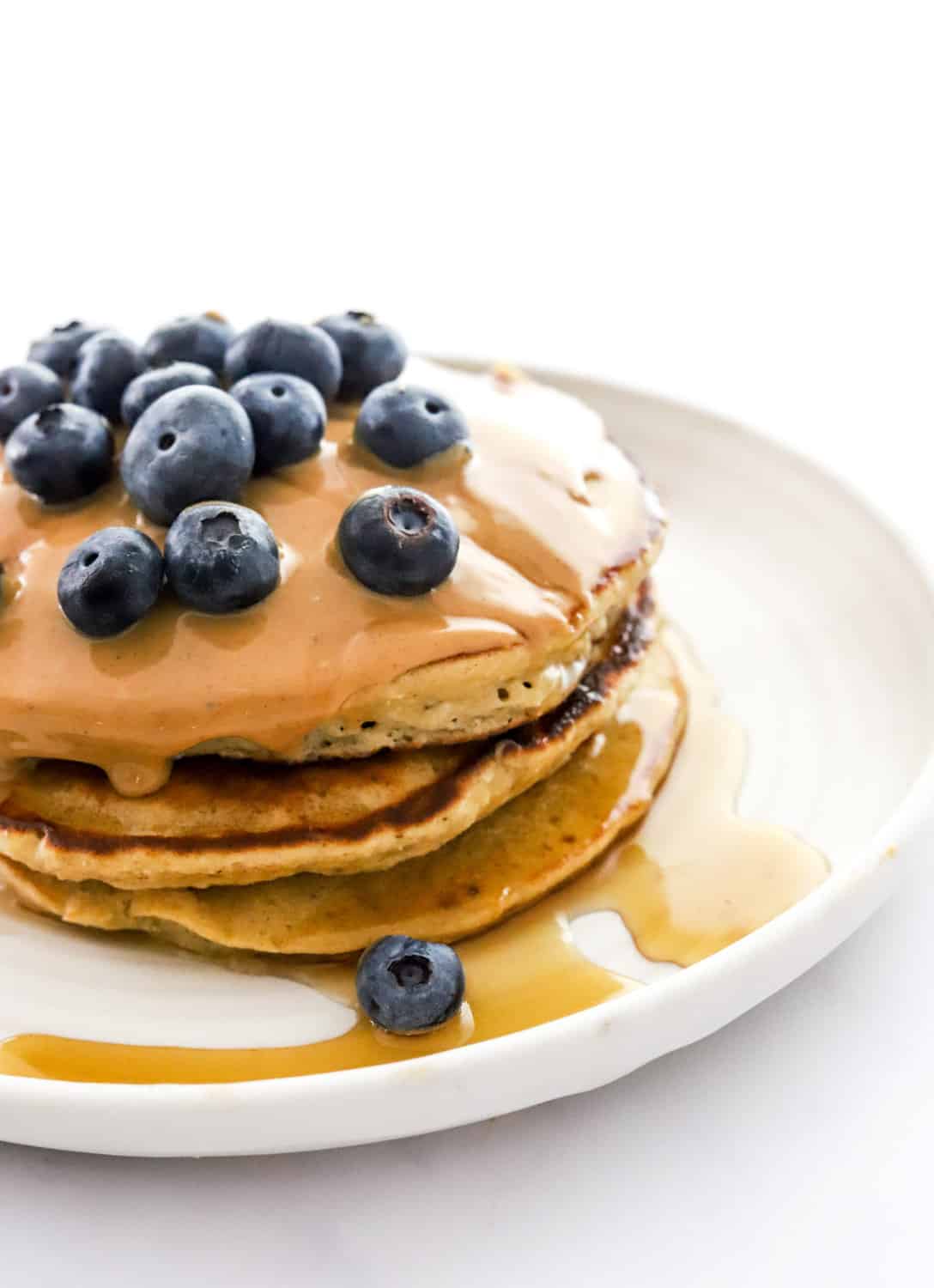 3 hot cakes stacked on of one another with syrup on them and topped with peanut butter and blueberries on a white plate. 
