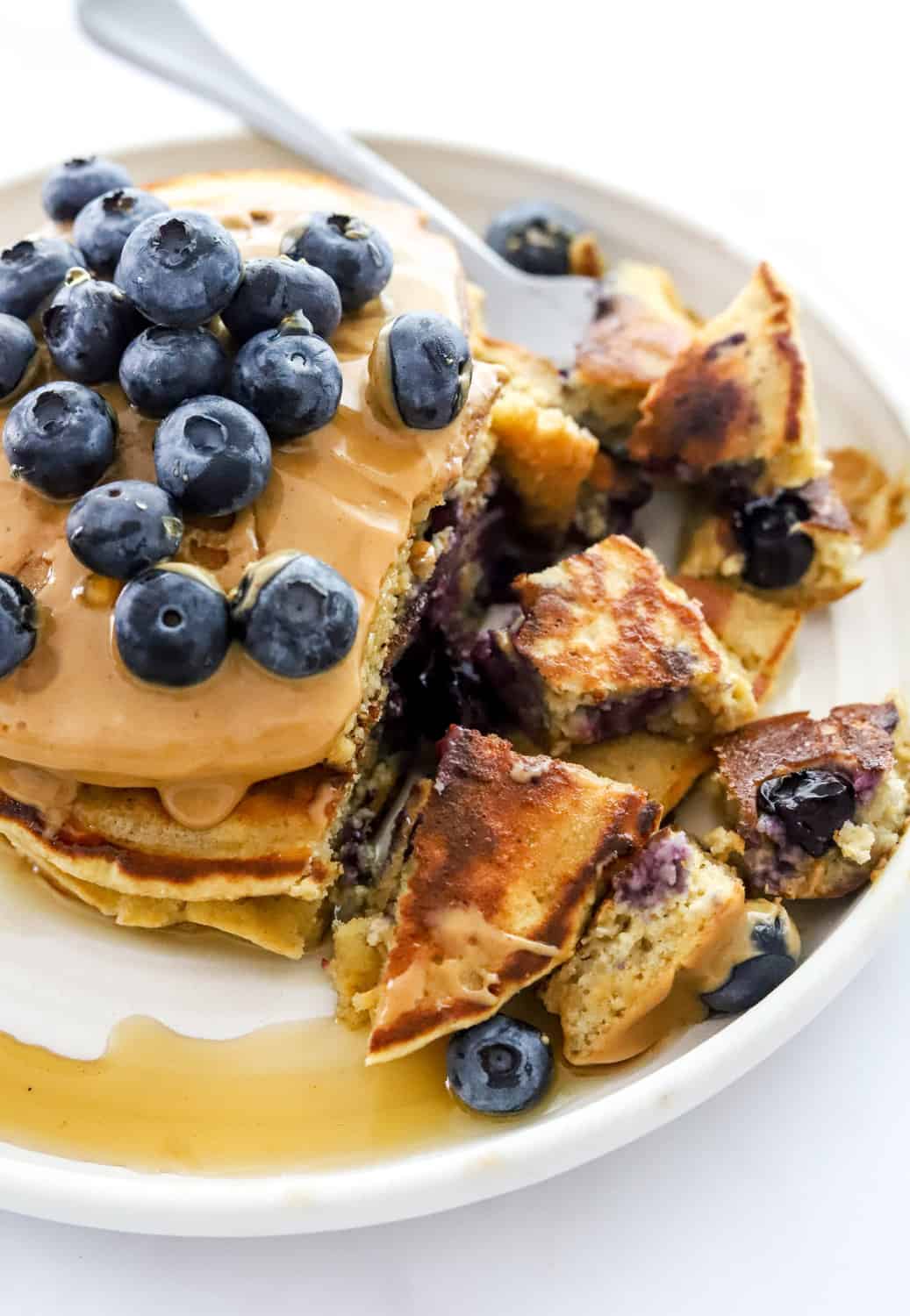 Stack of blueberry pancakes with peanut butter and blueberries on them with some of the pancakes cut up with a fork on the plate. 