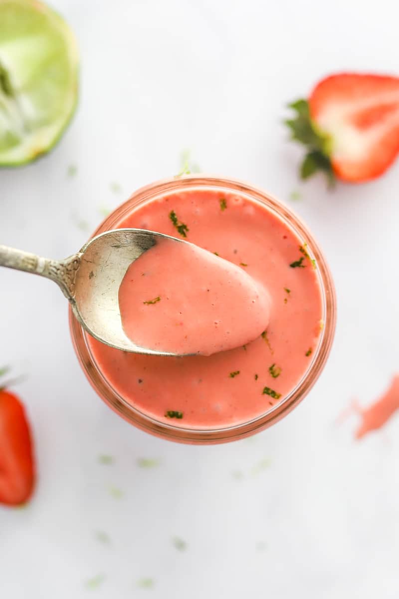 Spoon lifting strawberry vinaigrette dressing out of a glass jar with a sliced lime and a strawberry behind it and a strawberry in front of it. 