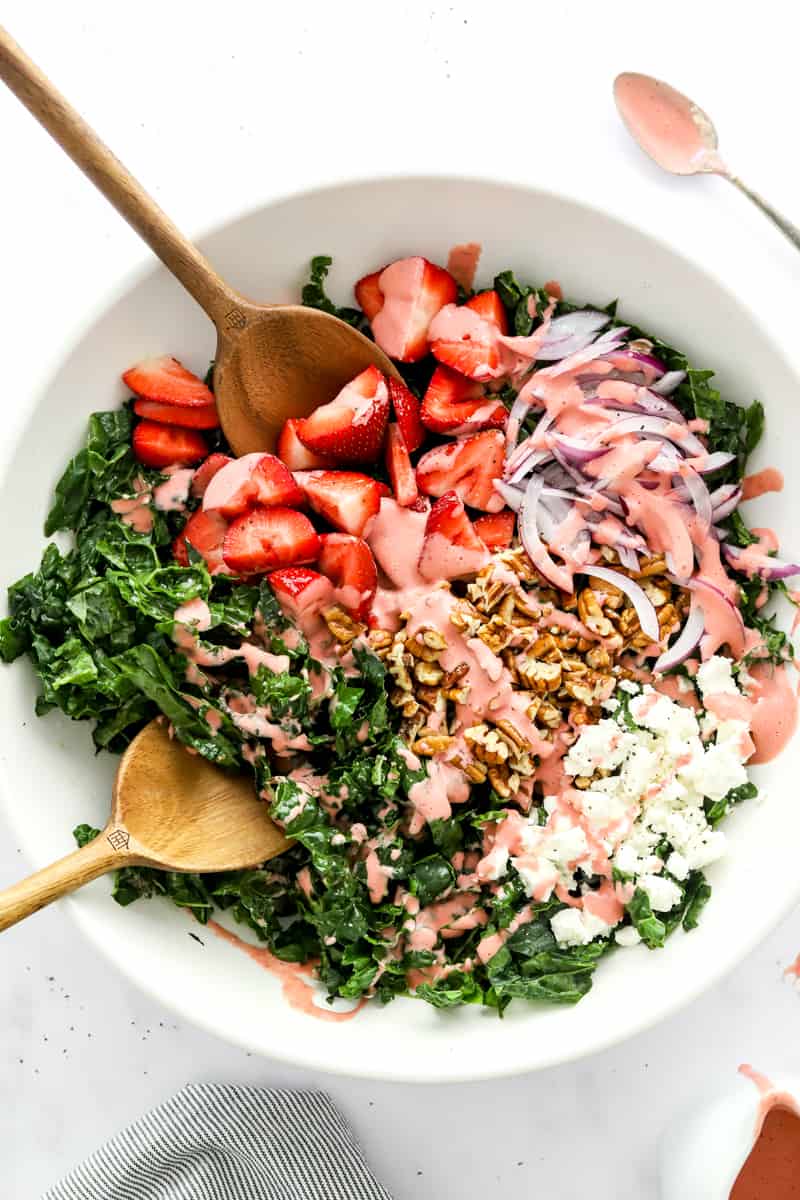 Strawberry kale salad in a white salad bowl topped with strawberry dressing with wooden serving spoons in the bowl. 