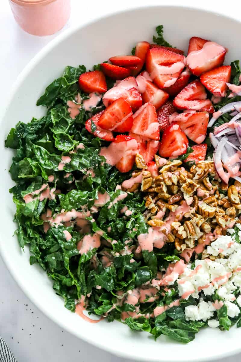 Close up of chopped kale salad with sliced strawberries, diced red onion, chopped nuts and a pink dressing drizzled on top in a large white bowl. 