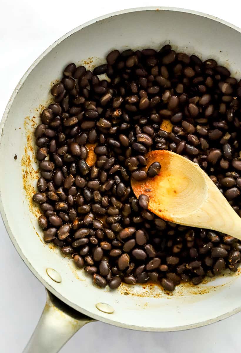 Black beans cooking in a skillet with a wooden spoon stirring them. 