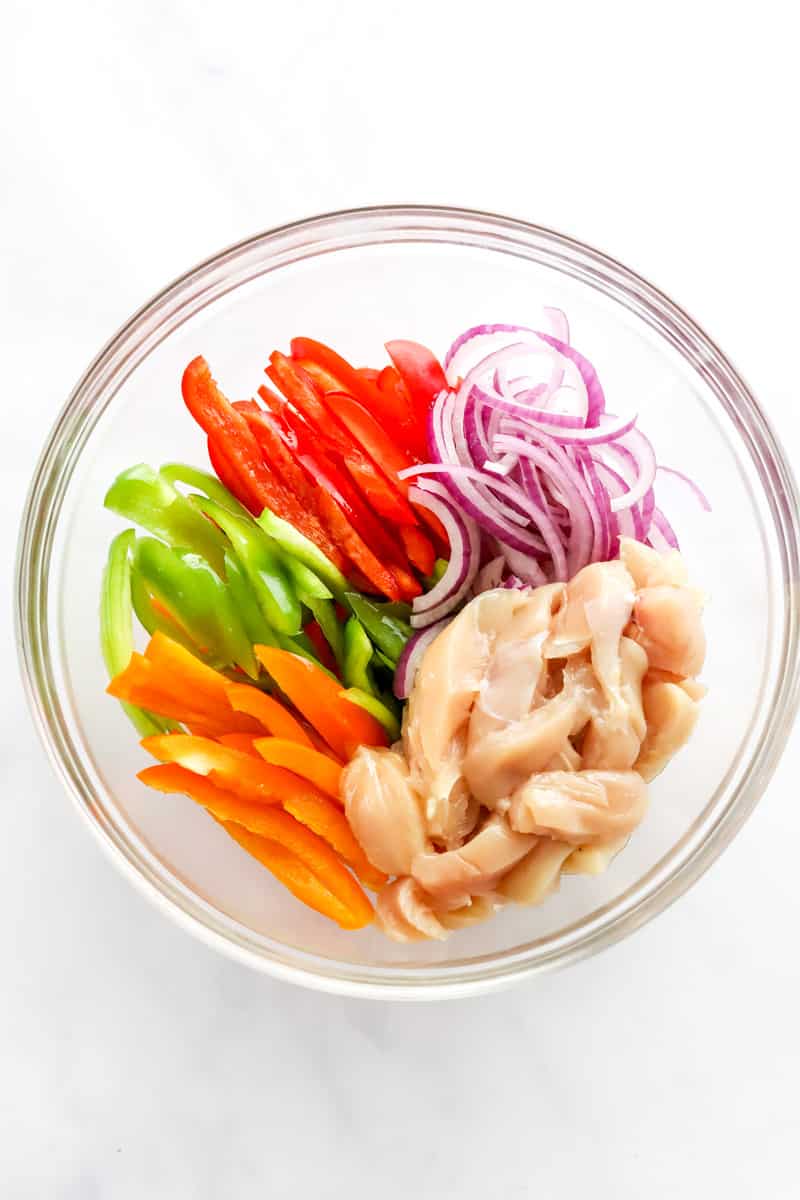 Uncooked sliced chicken breast, sliced peppers and onions in a glass mixing bowl. 