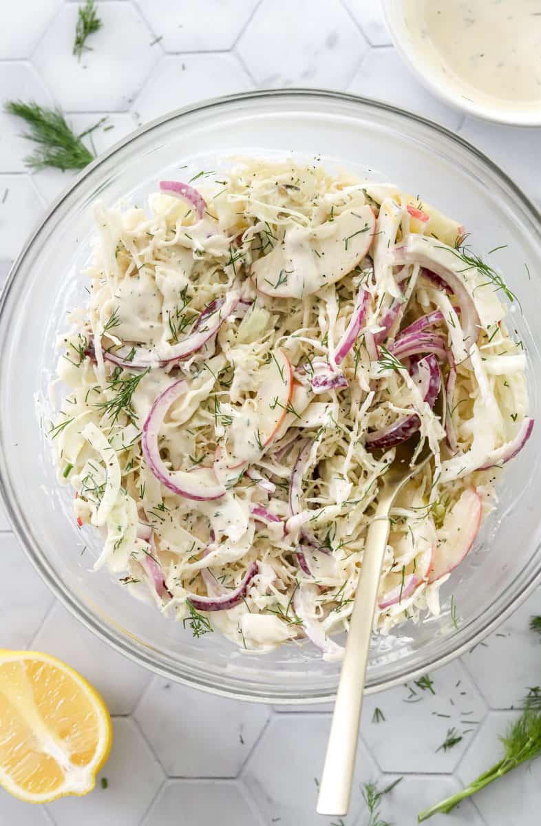 Coleslaw in a glass mixing bowl with a spoon in it with a slice of lemon in front of it and yogurt dressing behind it. 