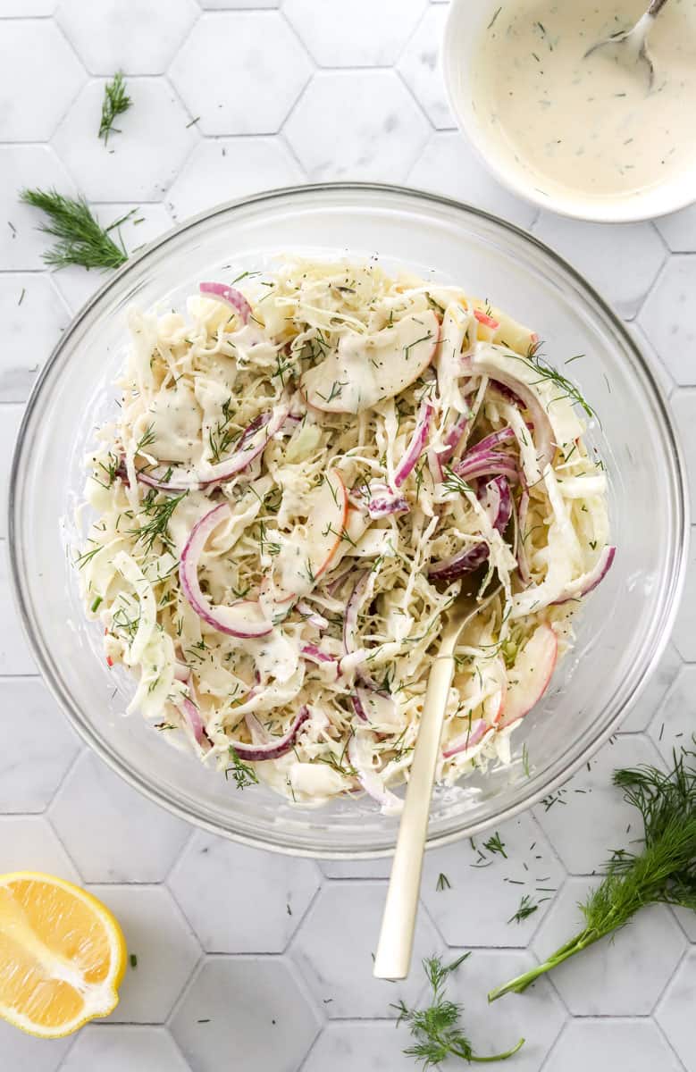 Creamy cabbage salad with sliced apple and red onion mixed up in a glass bowl with a spoon in it with dill and lemon in front of it. 
