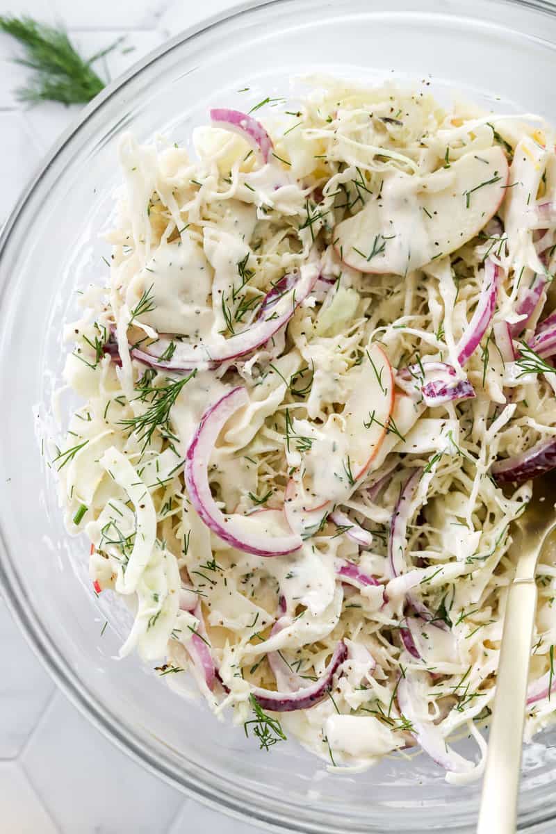 Sliced apple, fennel, cabbage and onion mixed in a creamy dressing in a glass bowl. 