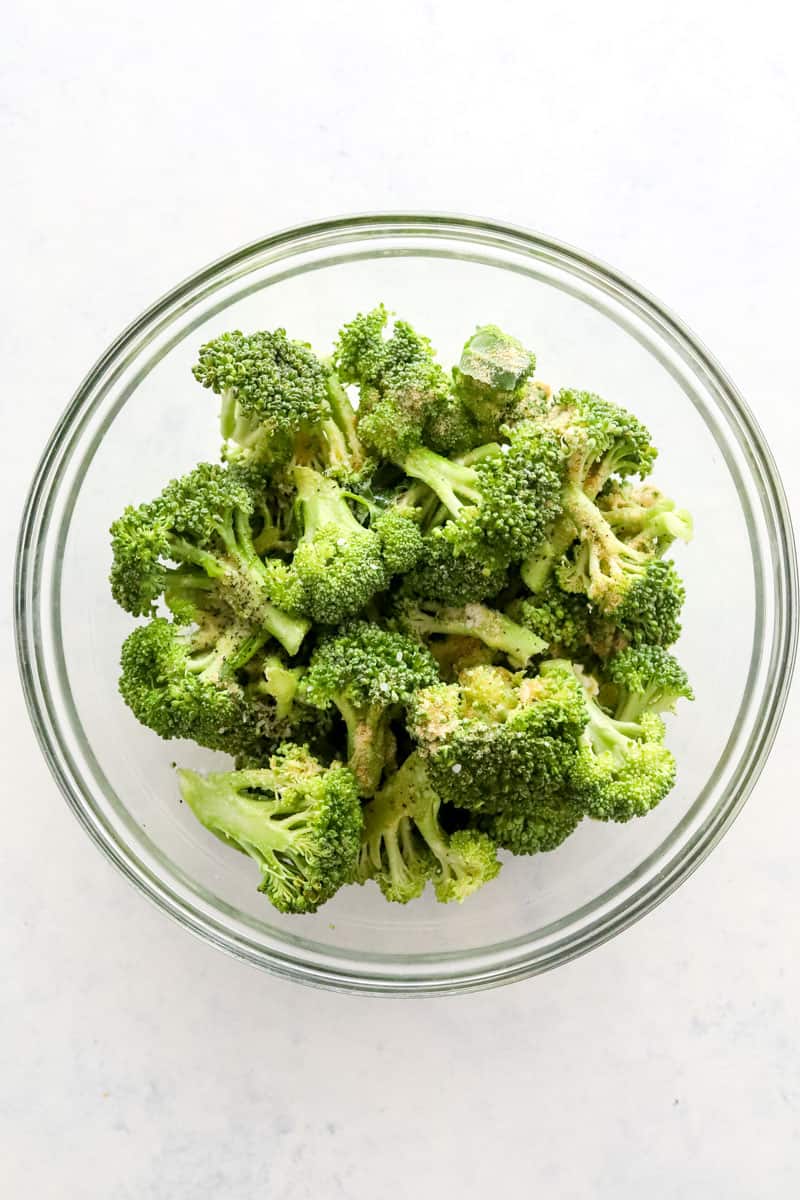Raw broccoli seasoned in a glass mixing bowl. 