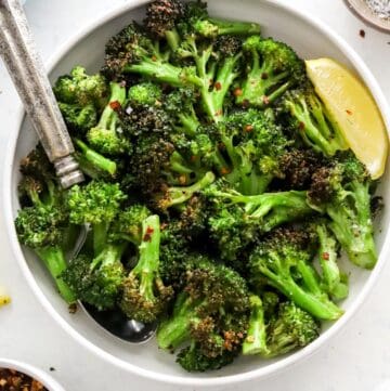 Air fryer broccoli in a serving bowl with a spoon in it with a lemon wedge next to it.