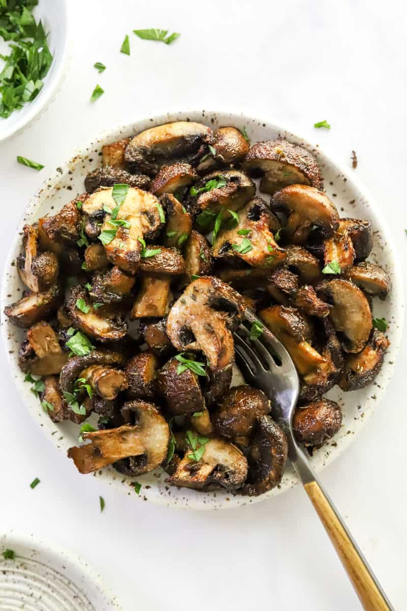 Golden cooked mushrooms in a bowl with a fork in the bowl with a bowl of herbs behind it. 