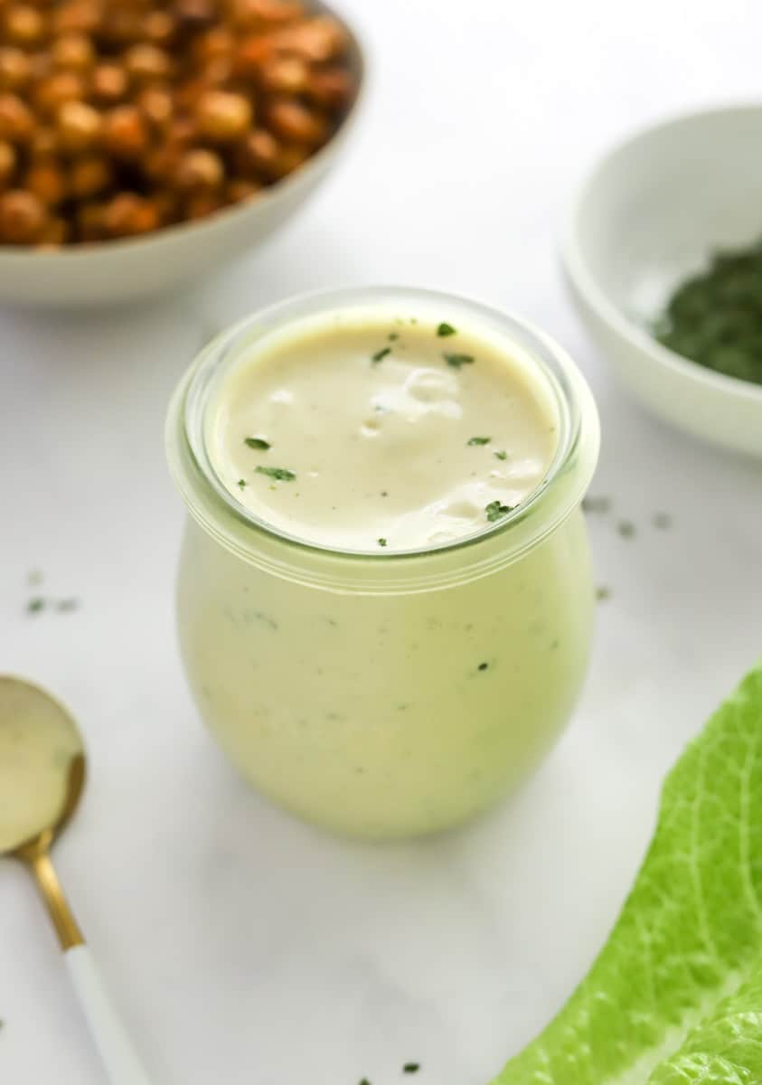 Healthy caesar dressing in a glass jar with chickpeas and herbs behind it. 