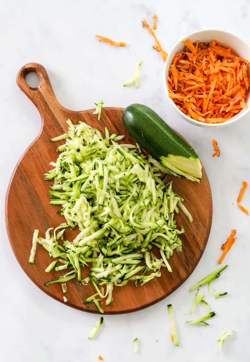 Shredded zucchini on a brown cutting board with a bowl of shredded sweet potato behind it. 