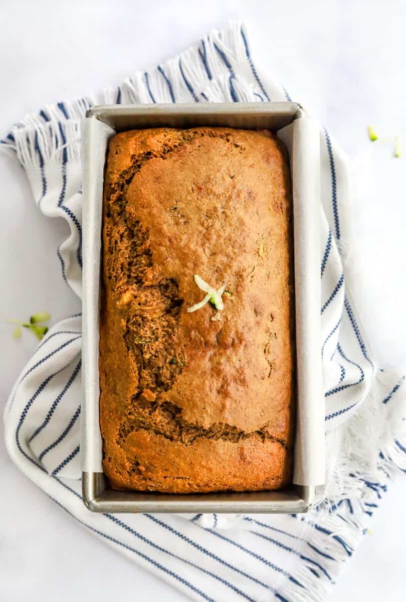 Baked golden brown loaf of zucchini bread in a silver loaf pan on top of a white and blue striped towel.