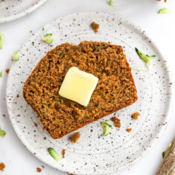 Piece of brown bread with a square pat of butter on it on a plate with more bread behind it.
