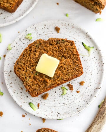 Piece of brown bread with a square pat of butter on it on a plate with more bread behind it.