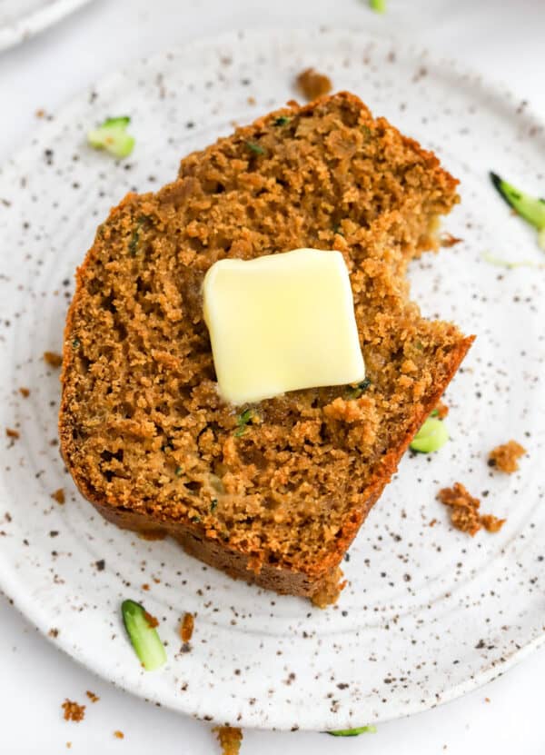 Close up of a pieces of banana bread with a pat of butter on it on a white plate with brown specs.