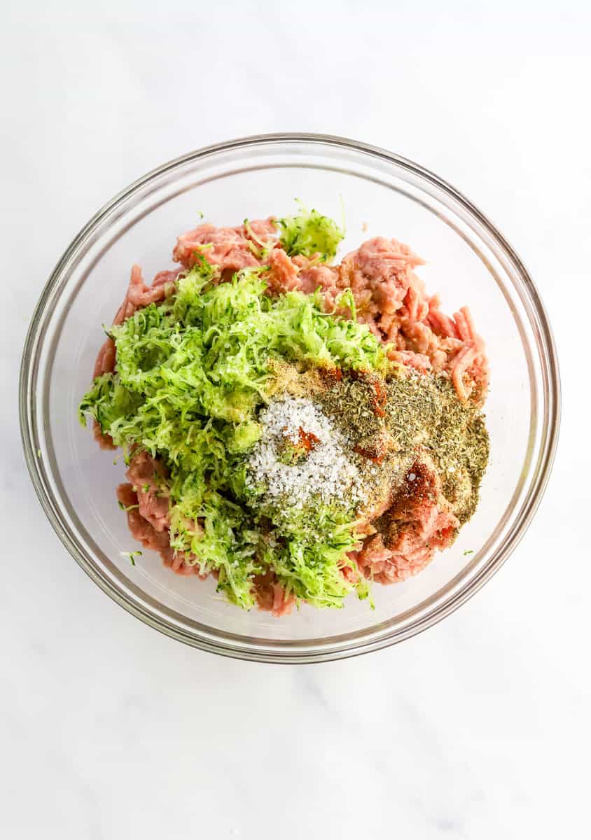 Raw ground turkey, shredded zucchini, oil and spices in a glass mixing bowl.
