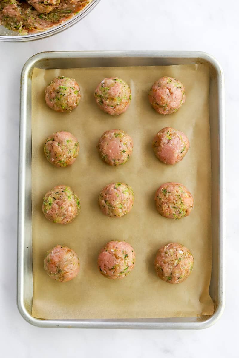 Rolled, uncooked meatballs on top of brown parchment paper on a silver baking sheet.