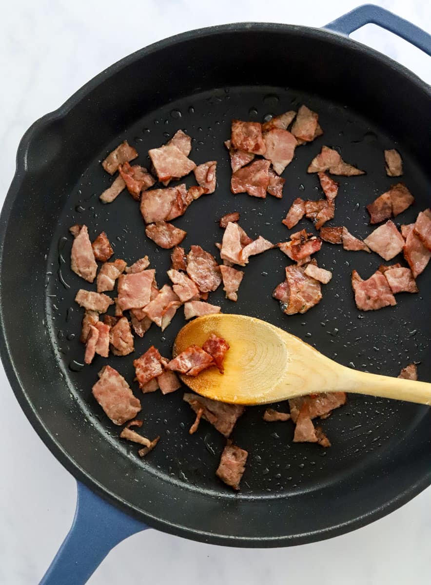Bacon cooking in a skillet with a wooden spoon stirring it. 