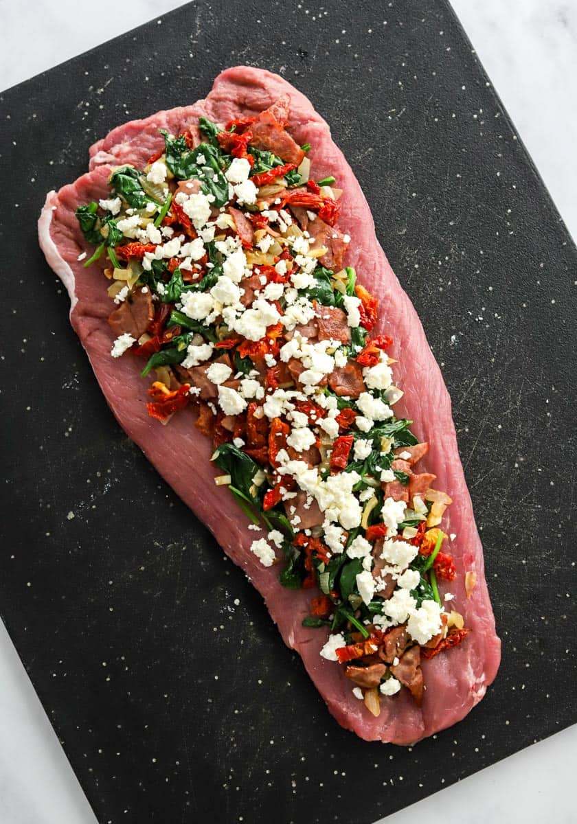 Flattended pork tenderloin on a black cutting board with spinach and feta filling bread out on it. 