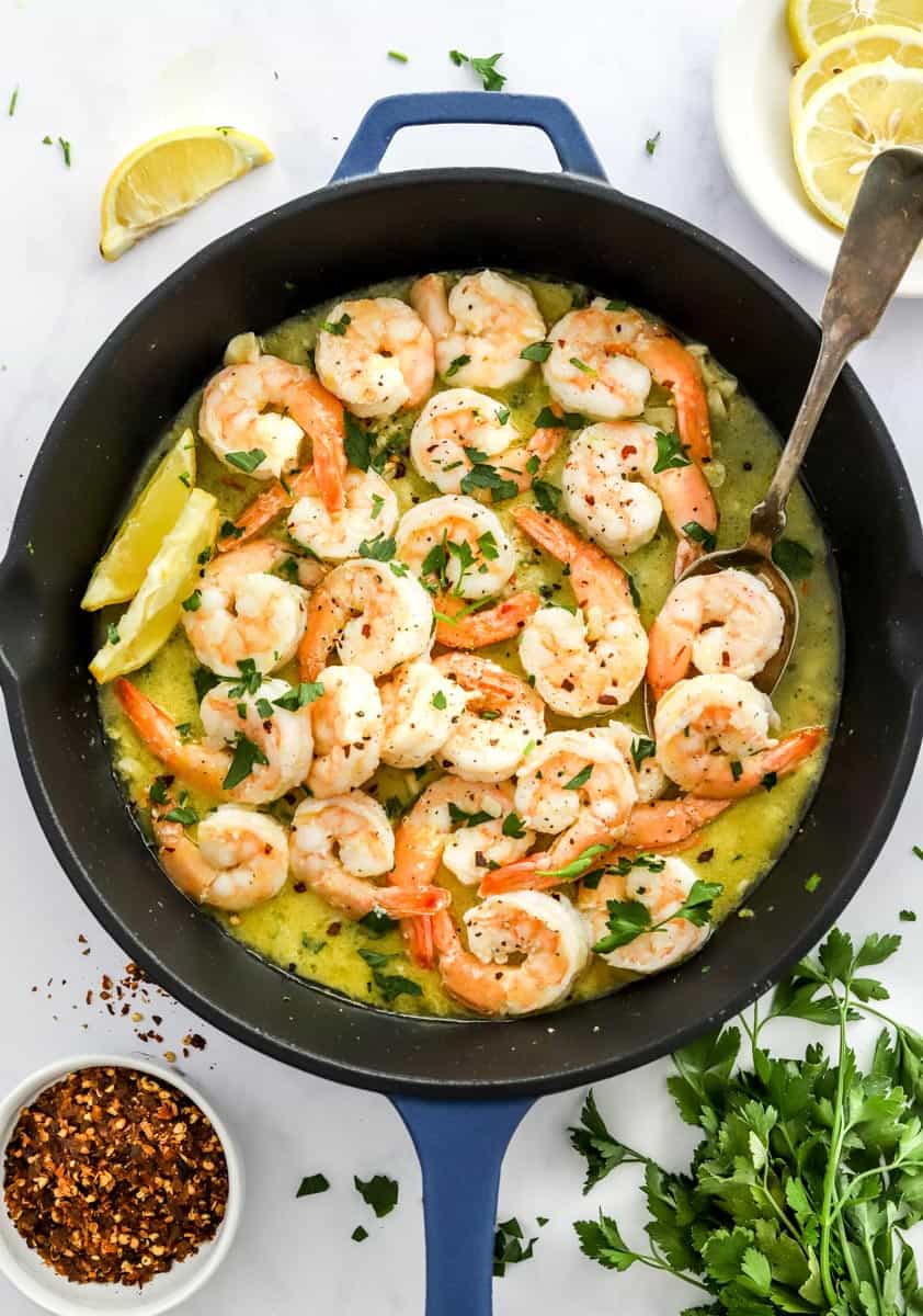 Cooked shrimp in a butter sauce in a black pan with lemon slices behind it and a bunch of fresh herbs in front of it. 
