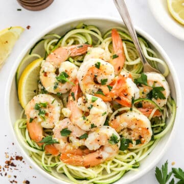Healthy shrimp scampi over zucchini noodles in a bow with sliced lemons and a pepper grinder behind it and red pepper flakes and herbs in front of it.