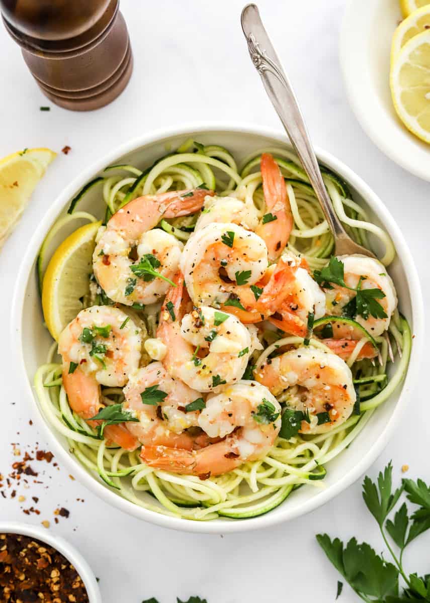 Healthy shrimp scampi over zucchini noodles in a bowl with sliced lemons and a pepper grinder behind it and red pepper flakes and herbs in front of it. 