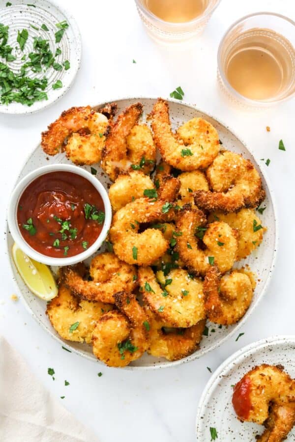 White plate filled with breaded shrimp with a bowl of red sauce next to it with a glass of wine behind it and a plate with another pieces of breaded shrimp in it in front of it.