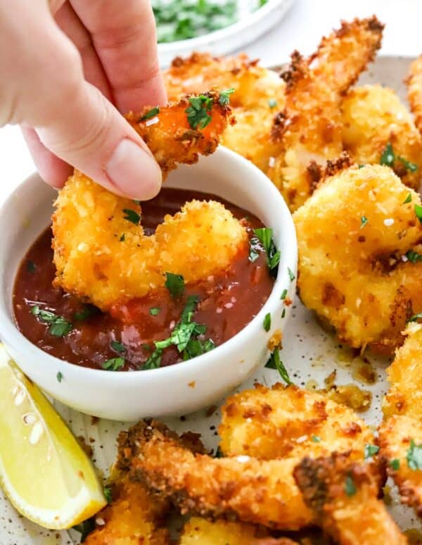 Hand dipping breaded coconut shrimp in a dish with cocktail sauce in it.