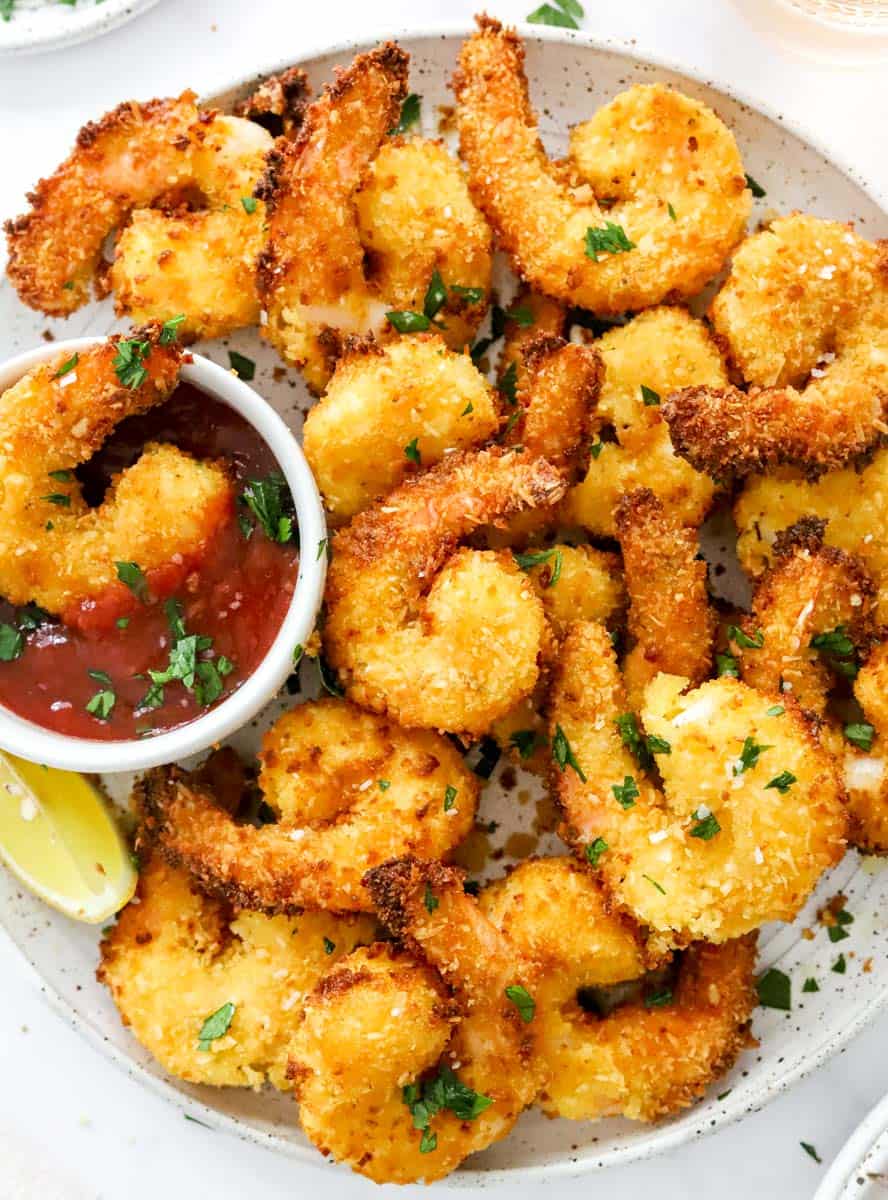 Crispy air fryer coconut shrimp on a plate with one of the shrimp in a bowl fo cocktail sauce. 
