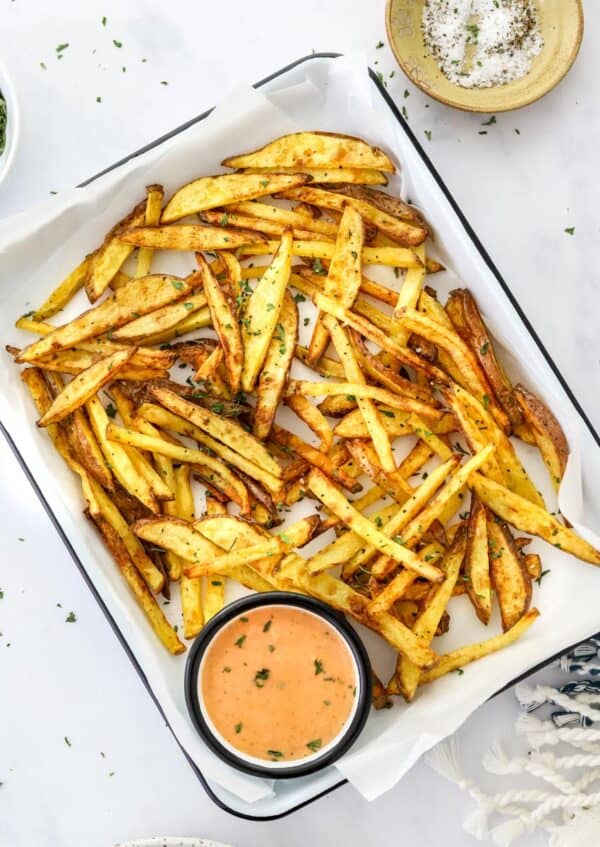 Crispy golden homemade French fries on a white dish with a dipping sauce with a yellow dish of salt and pepper behind it.