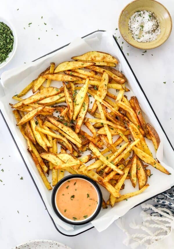 White platter with a black rim lined with white parchment paper with golden yellow fries on it with a bowl of salt and pepper and a bole of herbs behind it and a sipping sauce in a bowl on the platter with the fries.