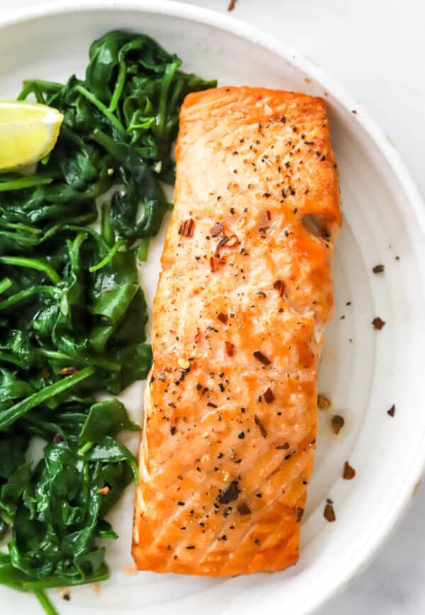 Close up shot of cooked piece of salmon with pepper on it next to some cooked spinach.
