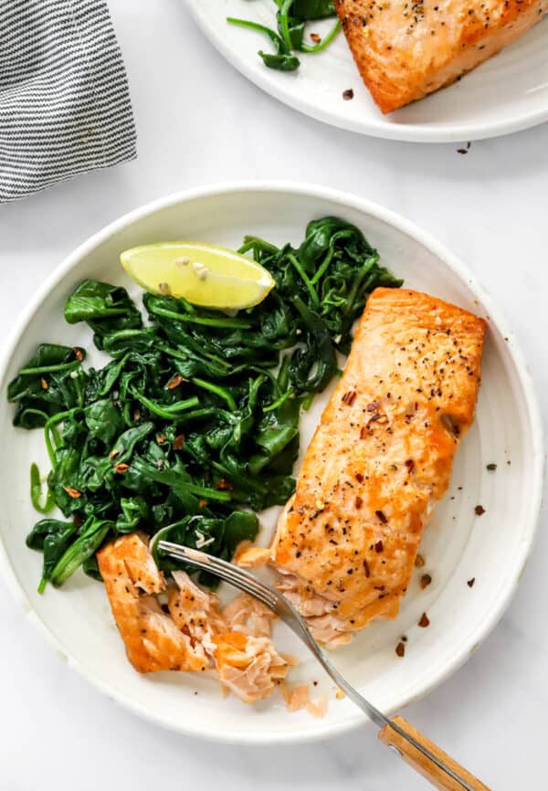 Cooked salmon filet on a round white plate next to sautéed greens with a another plate of salmon and a stripped towel behind it.