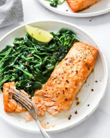 Plate with a cooked golden salmon filet on it next to spinach with another plate of salmon behind it.