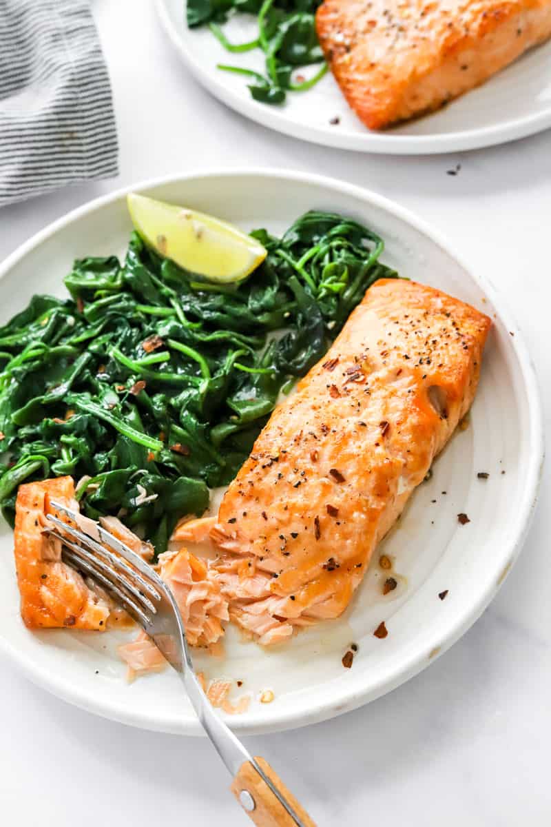 Plate with a cooked golden salmon filet on it next to spinach with another plate of salmon behind it.